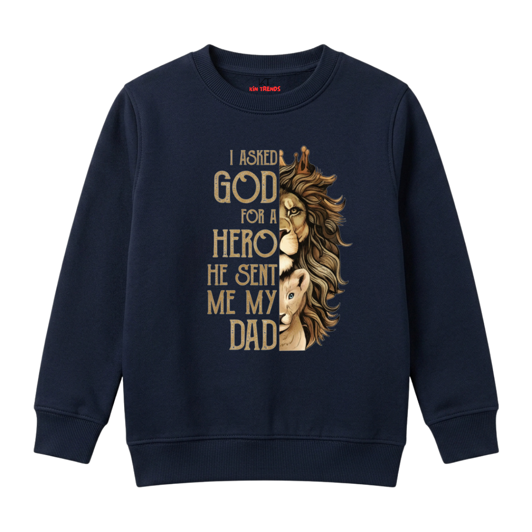 A navy blue crewneck sweatshirt featuring a graphic print of a lion half-severed from its human body, with the text 'I ASKED GOD FOR A HERO HE SENT ME MY DAD' in a gold serif font.
